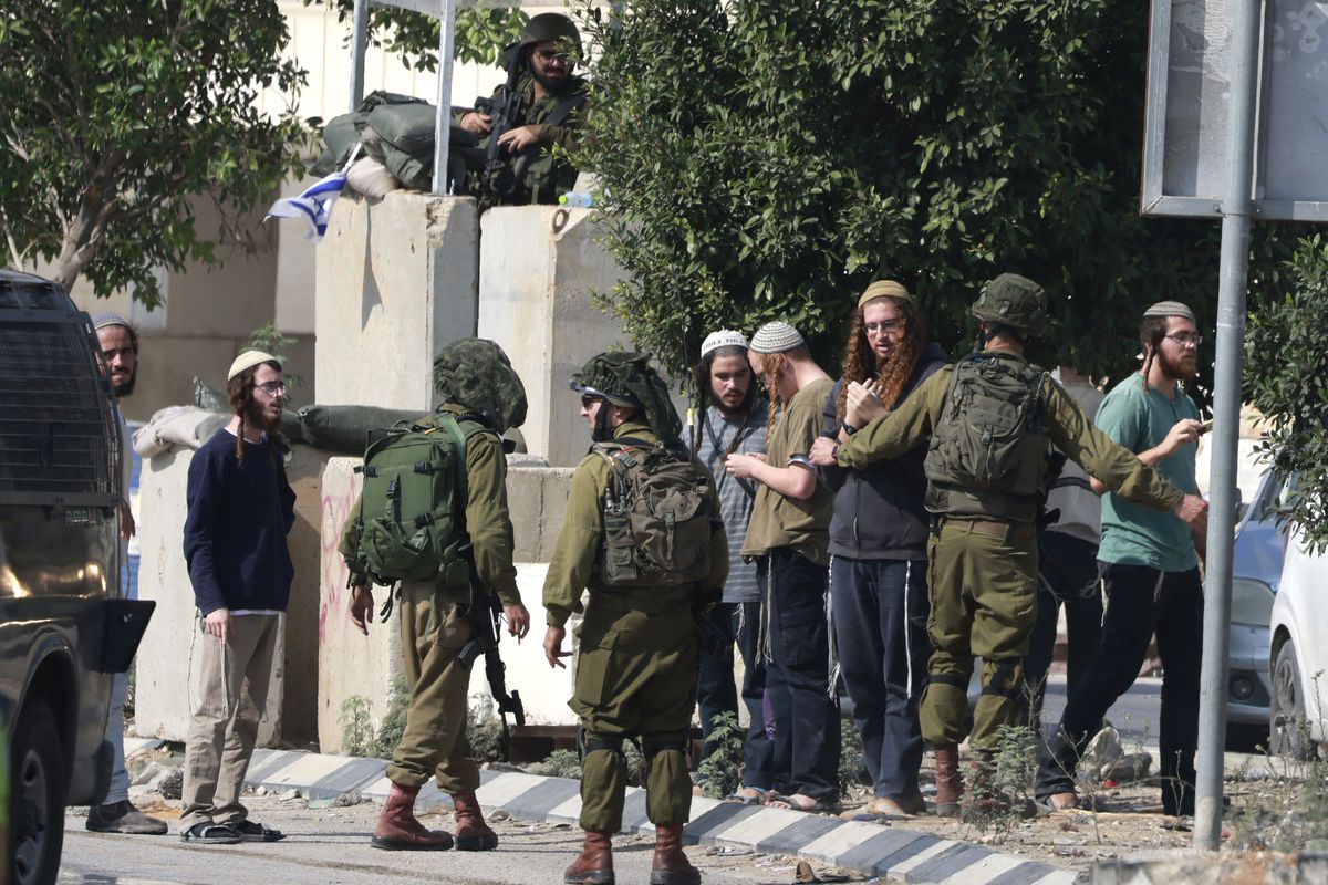 Israeli soldiers in uniforms walk with and push people out of a street, with others standing nearby watching.