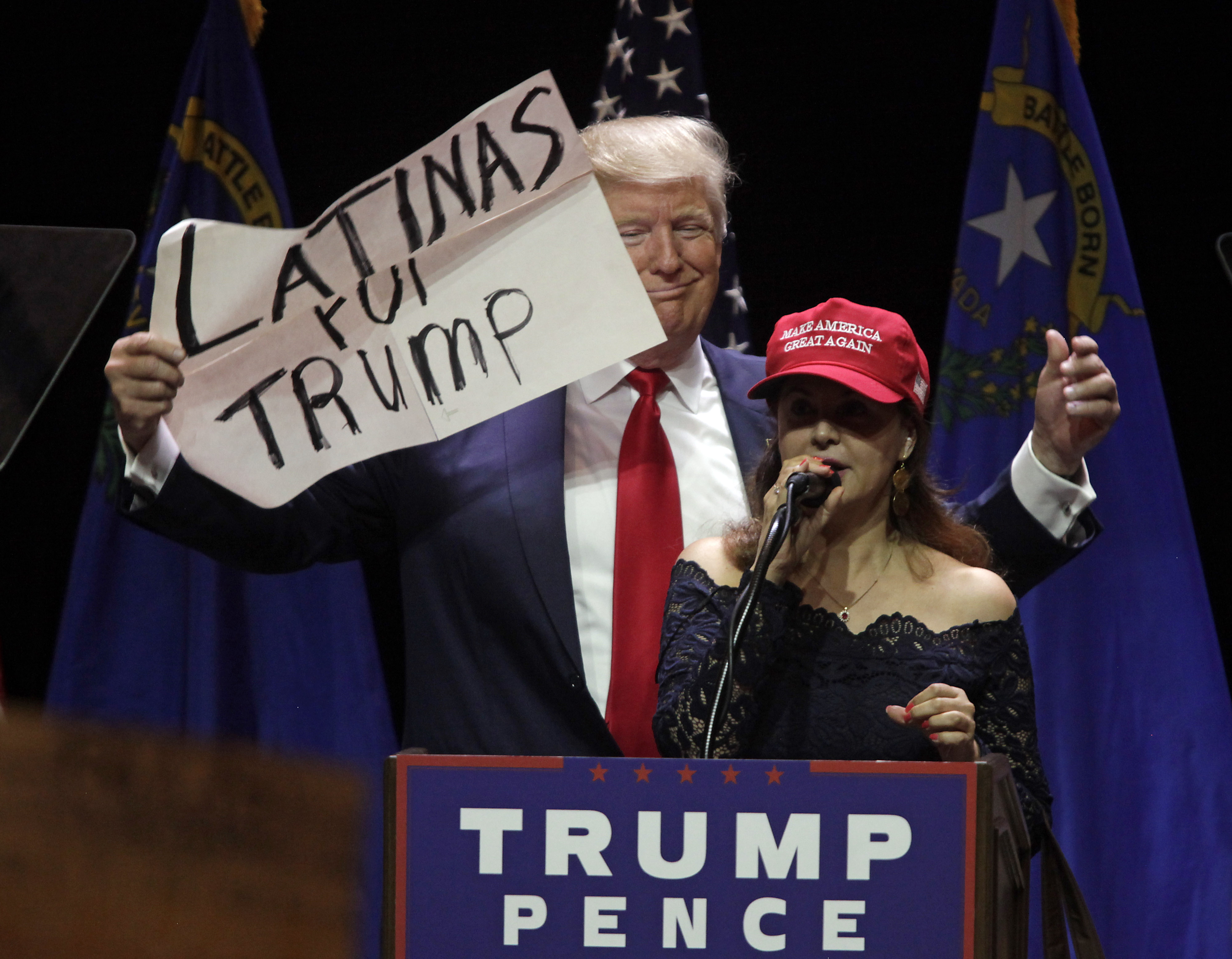 A woman in a MAGA hat speaks onstage while Donald Trump holds a sign behind her that reads “Latinas for Trump.”