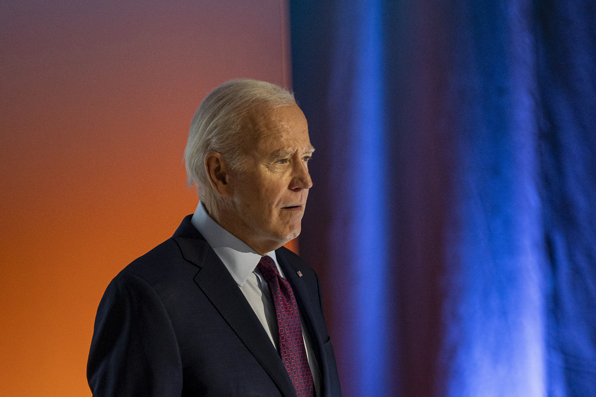US President Joe Biden standing in front of a blue curtain and orange background.