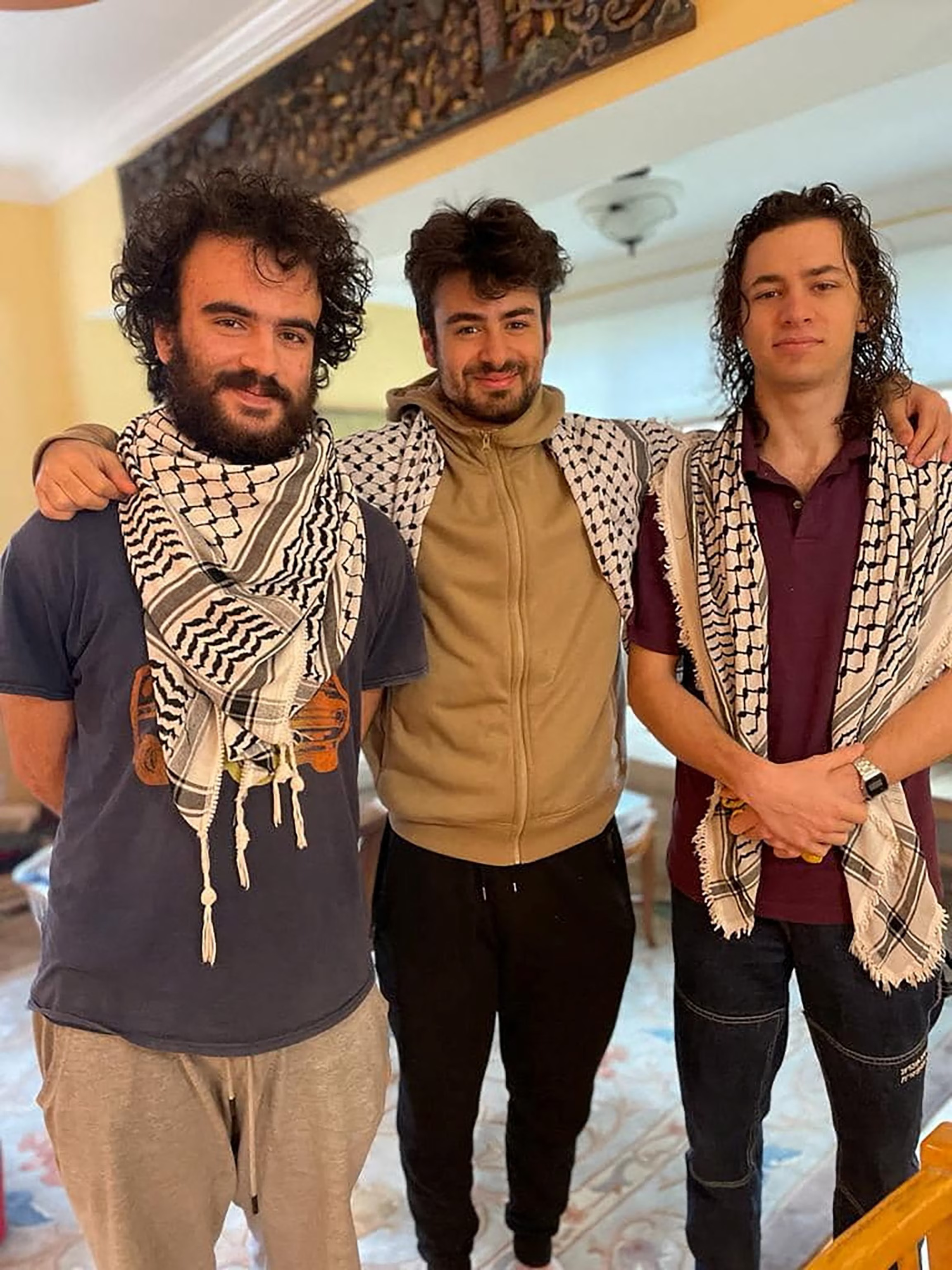 Three young men with dark hair and wearing keffiyeh black and white Palestinian scarves stand with their arms around each others’ shoulders.